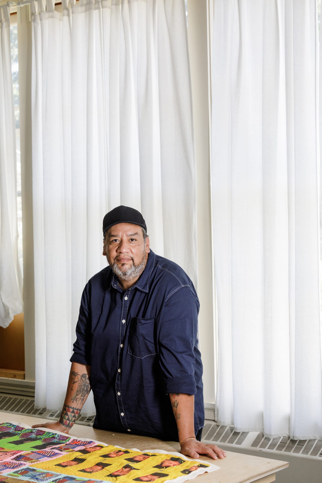 A man with wearing a denim shirt and a hat stands behind a table with printed fabric on top. Behind him are long white curtains on a rail