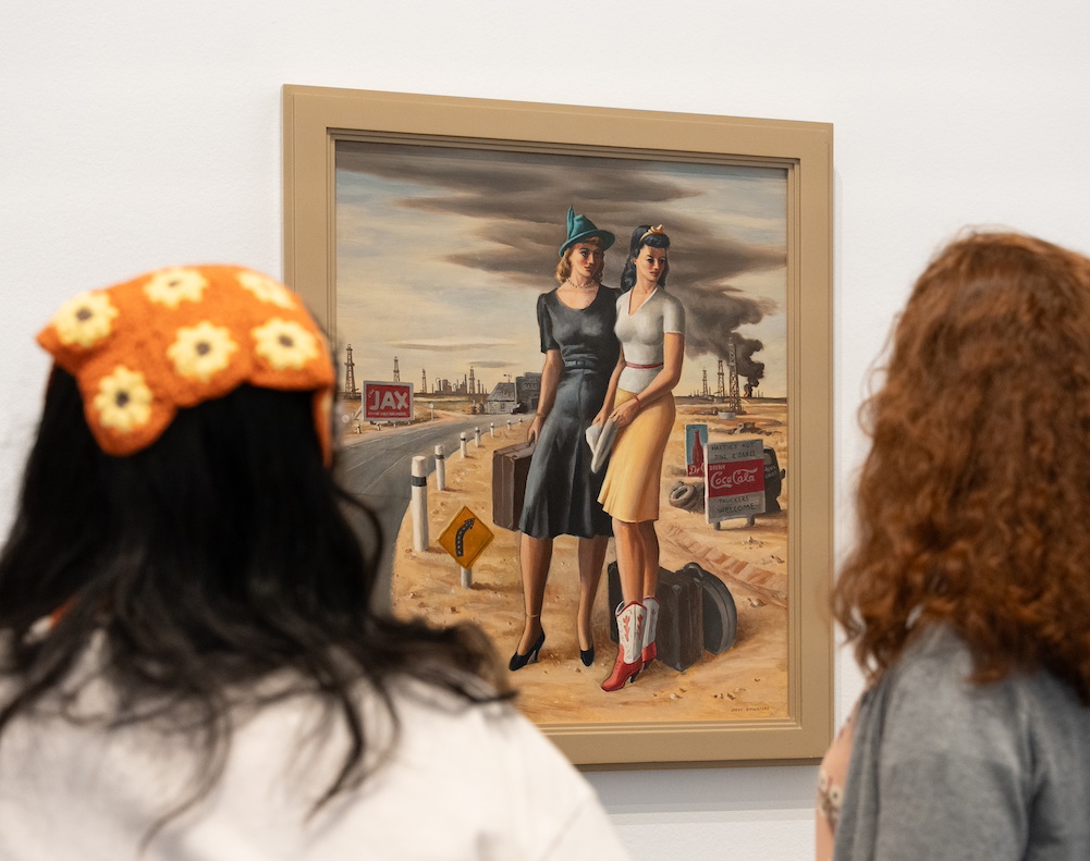 Two people looking at a painting of two women, one wearing cowgirl boots, standing in a desert area with what appears to be black smoke in the sky behind them
