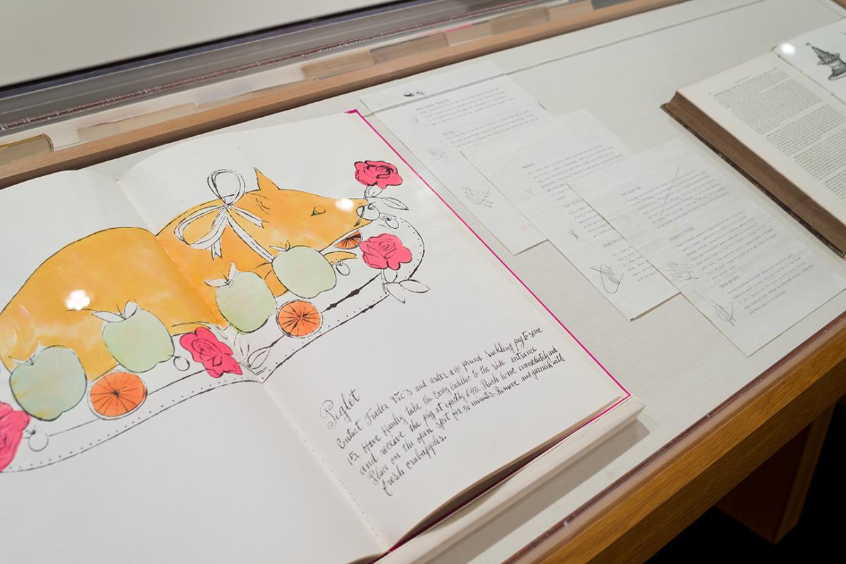Printed book propped open to feature an illustration of a roast pig on a plate with apples and a flower in it's mouth
