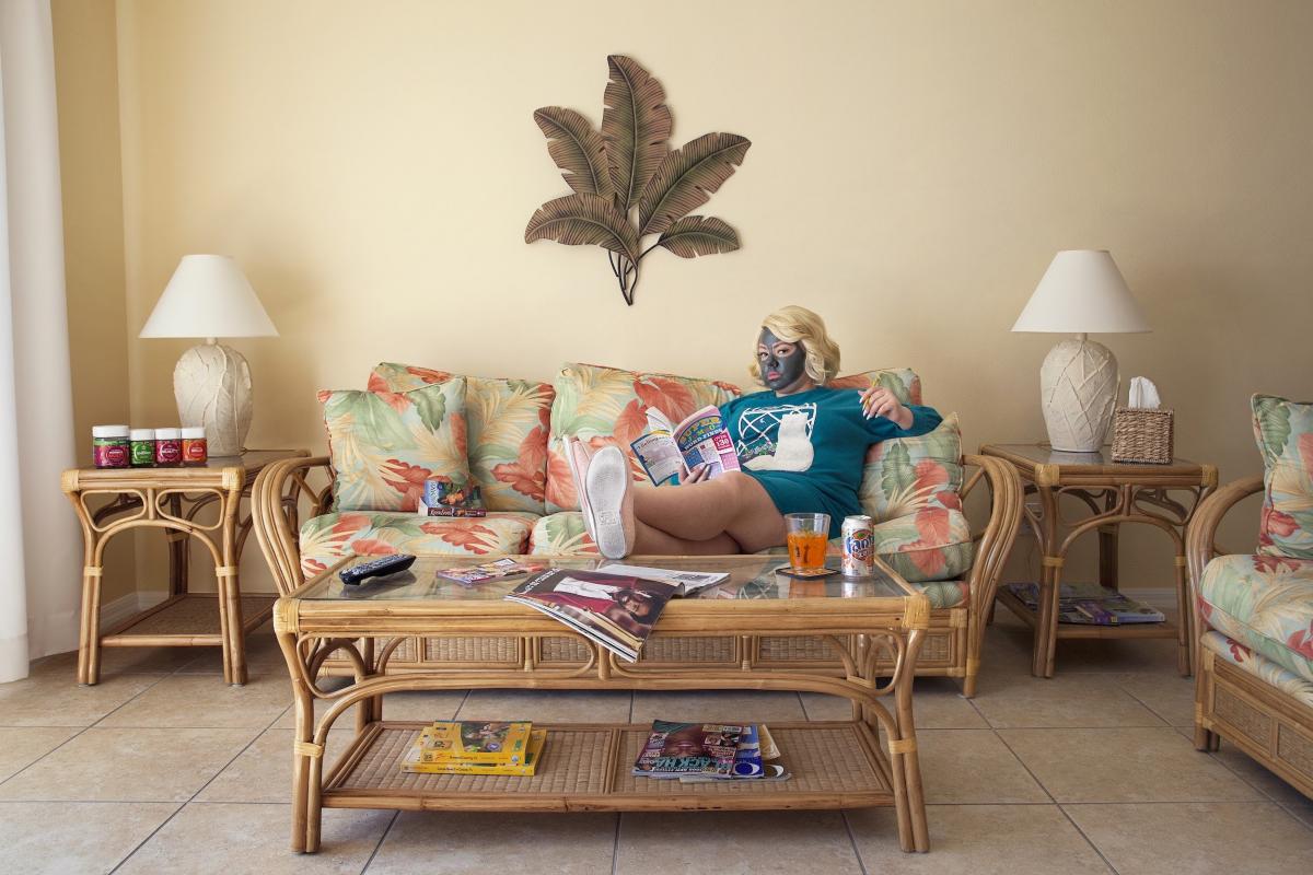 young woman sitting on a couch, her feet on the table before her; around her is more furniture and on the wall behind her a decoration made of large feathers